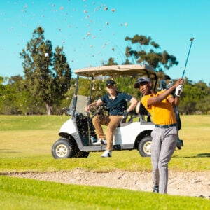 Caucasian young man sitting in cart looking at african american friend playing golf against sky